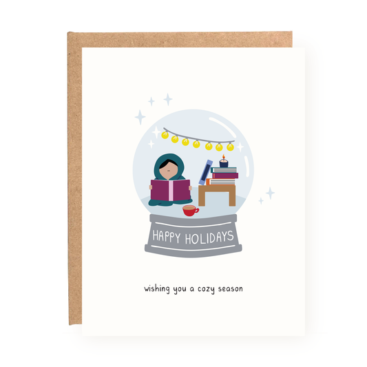 Cozy Snowglobe Holiday Card showing a person reading inside a snowglobe with books and holiday lights.