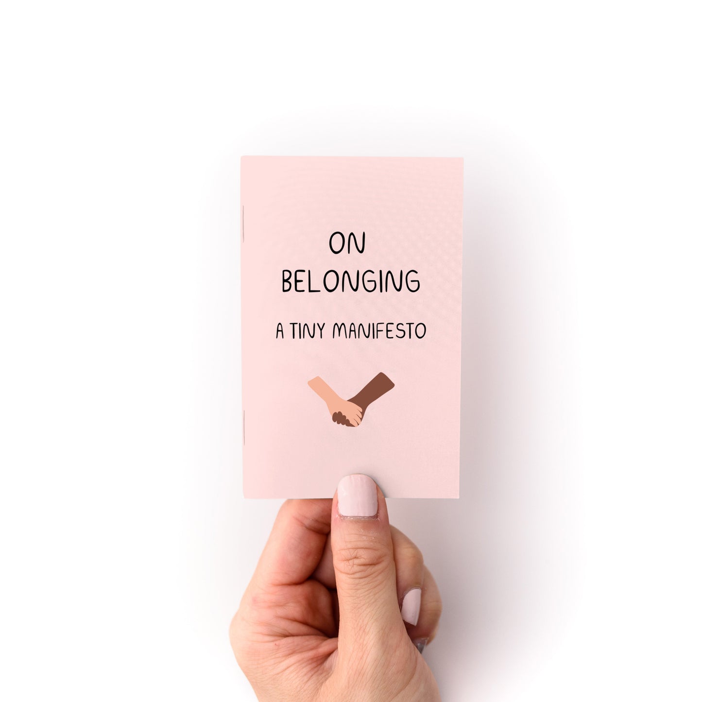 Hand holding On Belonging (A Tiny Manifesto) Zine, a small pink printed magazine about belonging with handshake illustration.