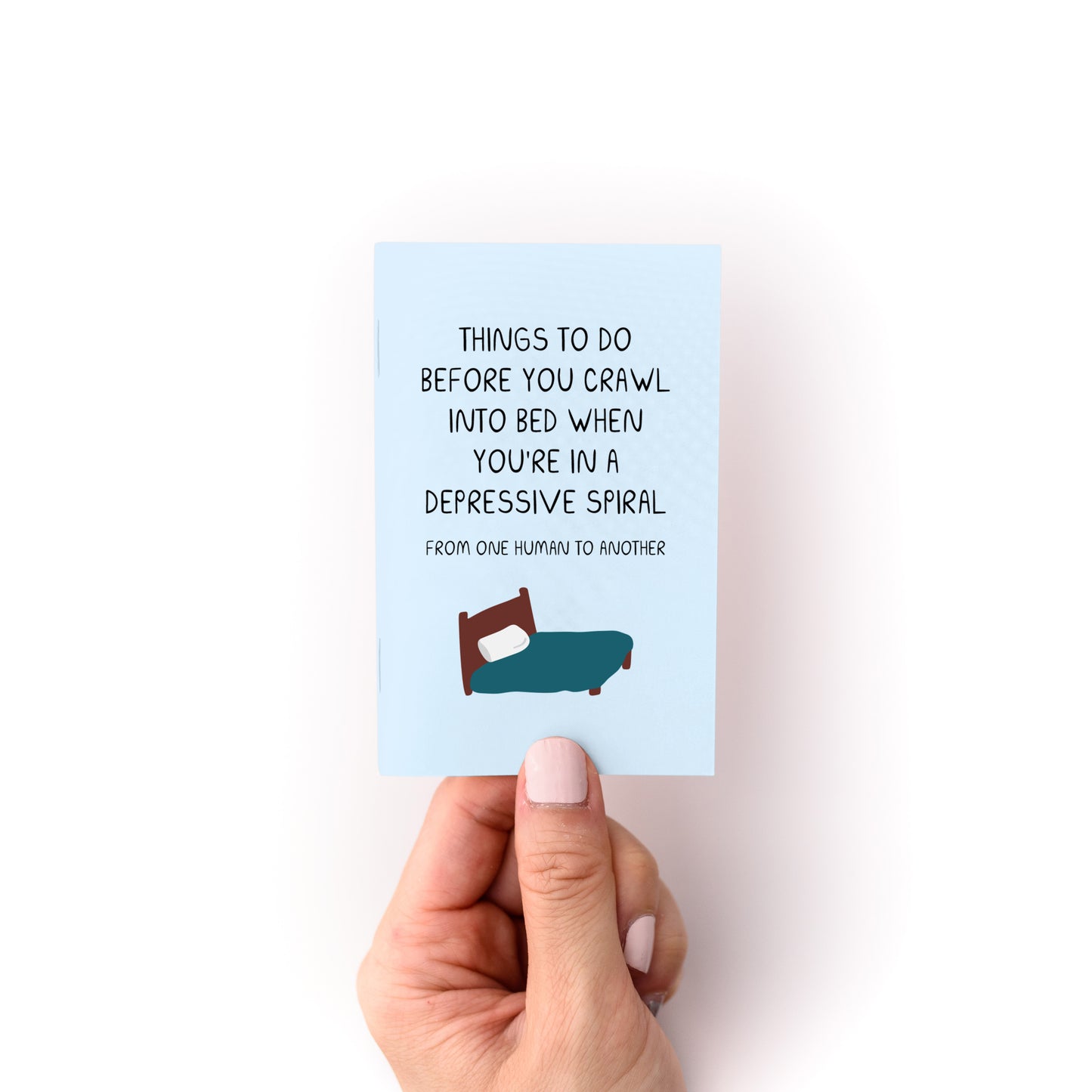 Hand holding Things to Do When You're in a Depressive Spiral Zine with bed illustration on blue cover.