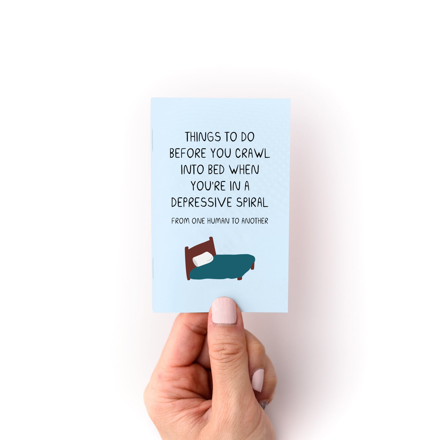 Hand holding Things to Do When You're in a Depressive Spiral Zine with bed illustration on blue cover.