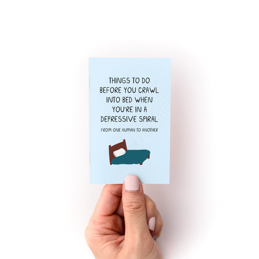 Hand holding Things to Do When You're in a Depressive Spiral Zine with bed illustration on blue cover.