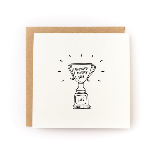 Thoughtful sympathy card or encouragement card for a survivor or anyone who is going through a tough time. The card has a white background and black illustration of a trophy that says, Survived Another Year.