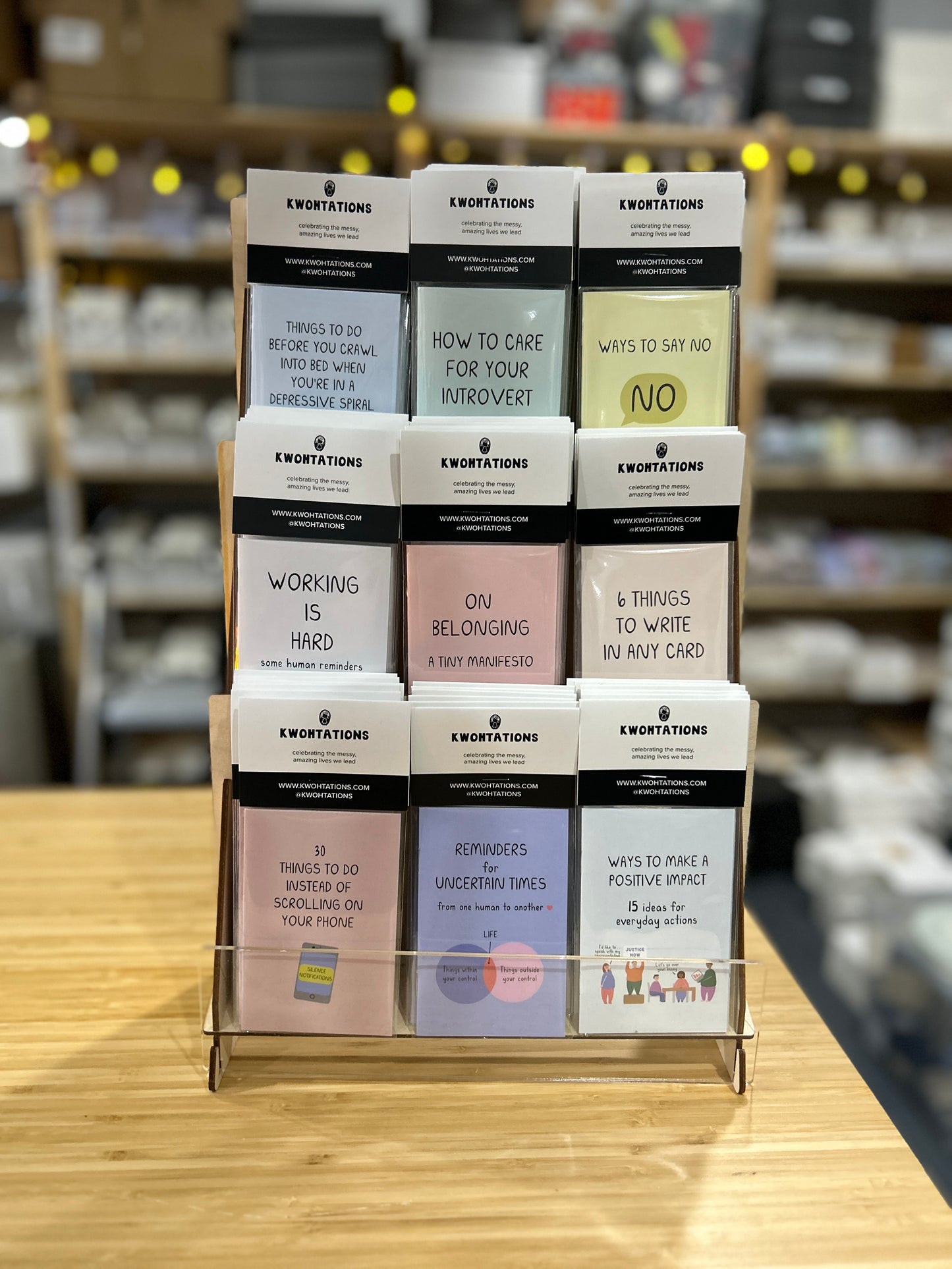 Wooden display rack holding 54 colorful Kwnotations zines with thoughtful titles arranged in three rows on wooden table