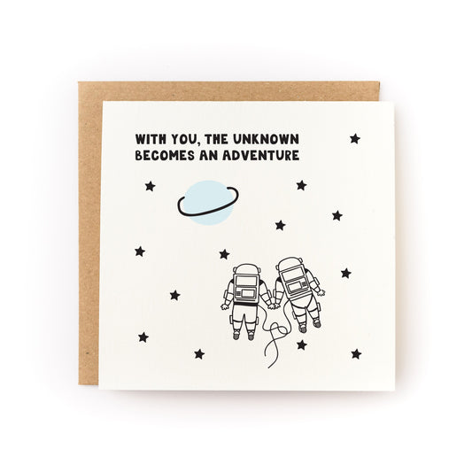 With You the Unknown Becomes an Adventure Letterpress Card with two astronauts holding hands among stars and a planet.