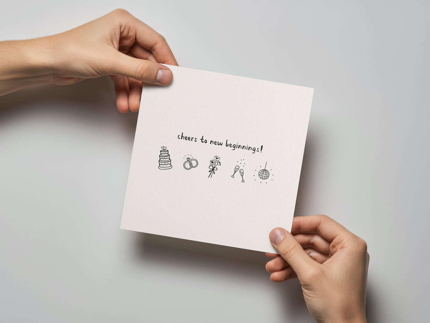 White square wedding card held by hands with black line illustrations and text "cheers to new beginnings!" on front