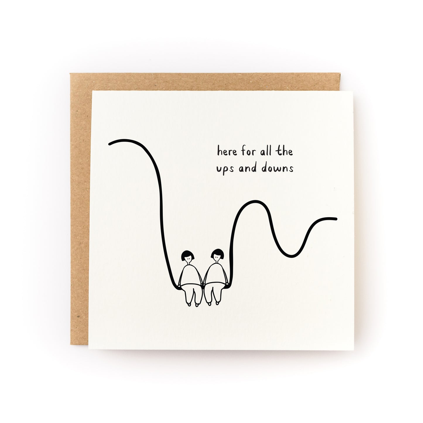 Square white greeting card featuring a minimalist black line drawing of two people holding hands while sitting on a curved line, with the handwritten text "here for all the ups and downs," paired with a brown kraft envelope.