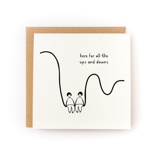 Square white greeting card featuring a minimalist black line drawing of two people holding hands while sitting on a curved line, with the handwritten text "here for all the ups and downs," paired with a brown kraft envelope.