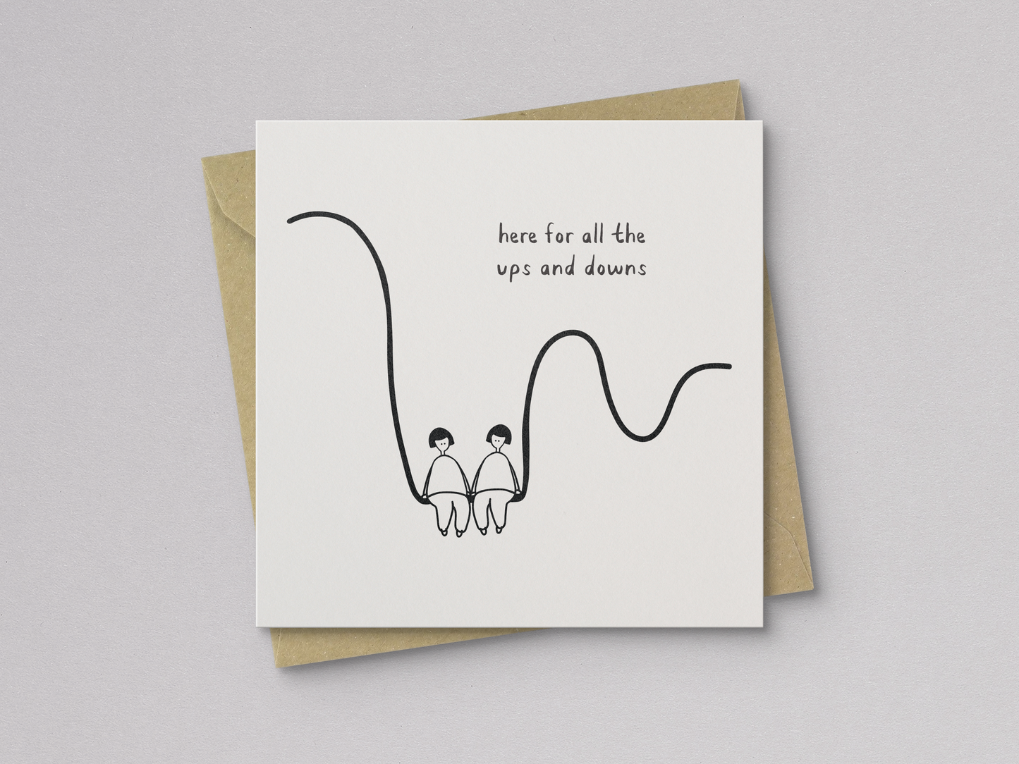 Square white greeting card with a minimal black line drawing of two people sitting side by side on a wavy line, accompanied by handwritten text reading "here for all the ups and downs," paired with a brown kraft envelope.