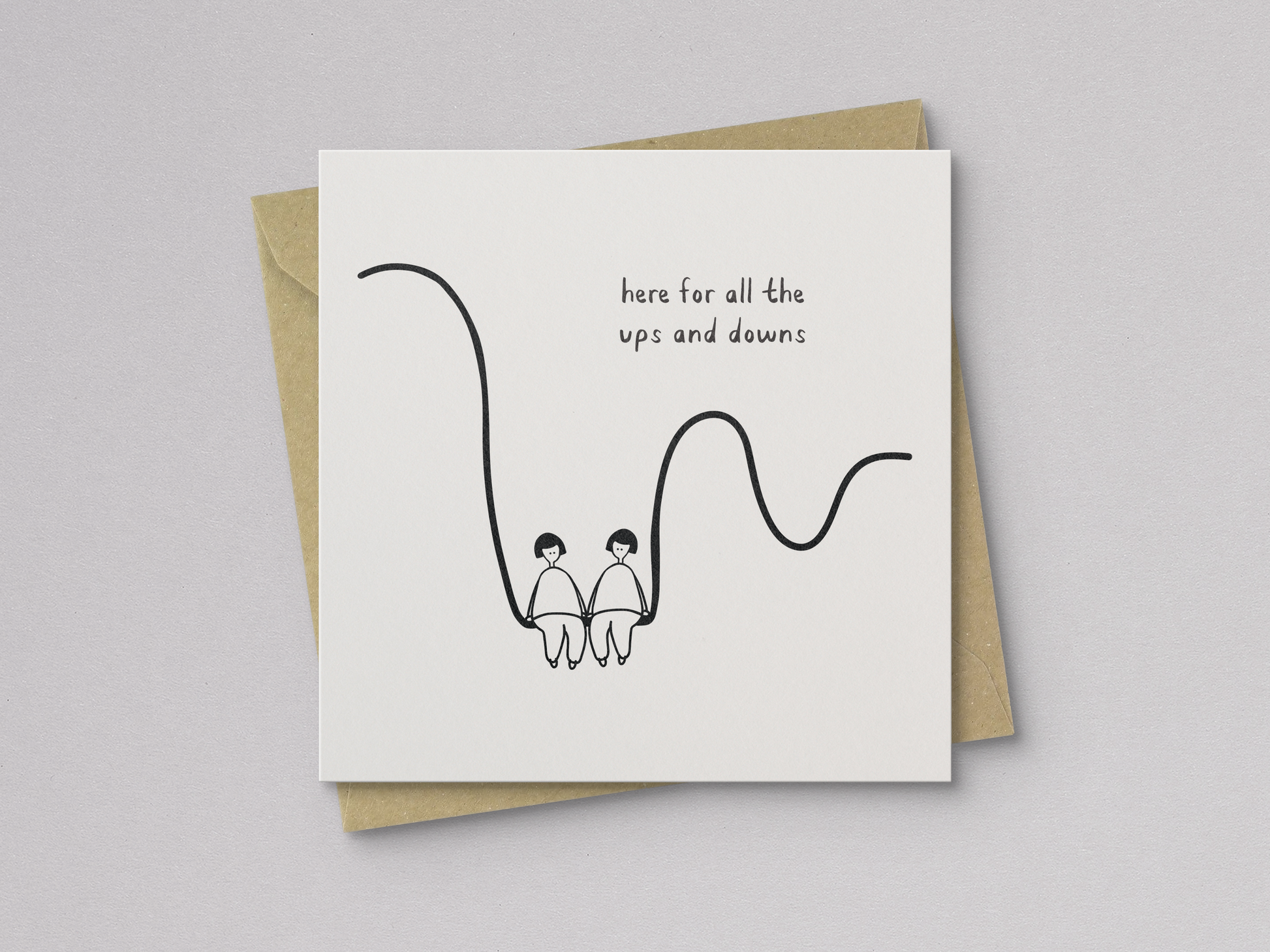 Square white greeting card with a minimal black line drawing of two people sitting side by side on a wavy line, accompanied by handwritten text reading "here for all the ups and downs," paired with a brown kraft envelope.
