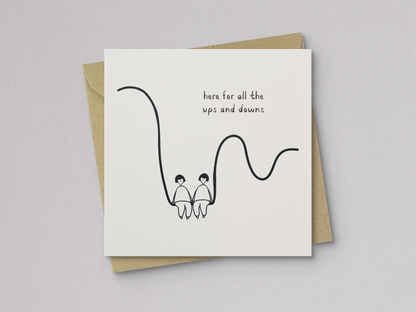 Square white greeting card with a minimal black line drawing of two people sitting side by side on a wavy line, accompanied by handwritten text reading "here for all the ups and downs," paired with a brown kraft envelope.