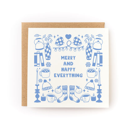 Square greeting card with light blue illustrations of cozy winter items including mittens, mugs, candles, books, hats, socks, and vases, surrounding the text "Merry and Happy Everything," paired with a brown kraft envelope.