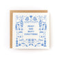Square greeting card with light blue illustrations of cozy winter items including mittens, mugs, candles, books, hats, socks, and vases, surrounding the text "Merry and Happy Everything," paired with a brown kraft envelope.