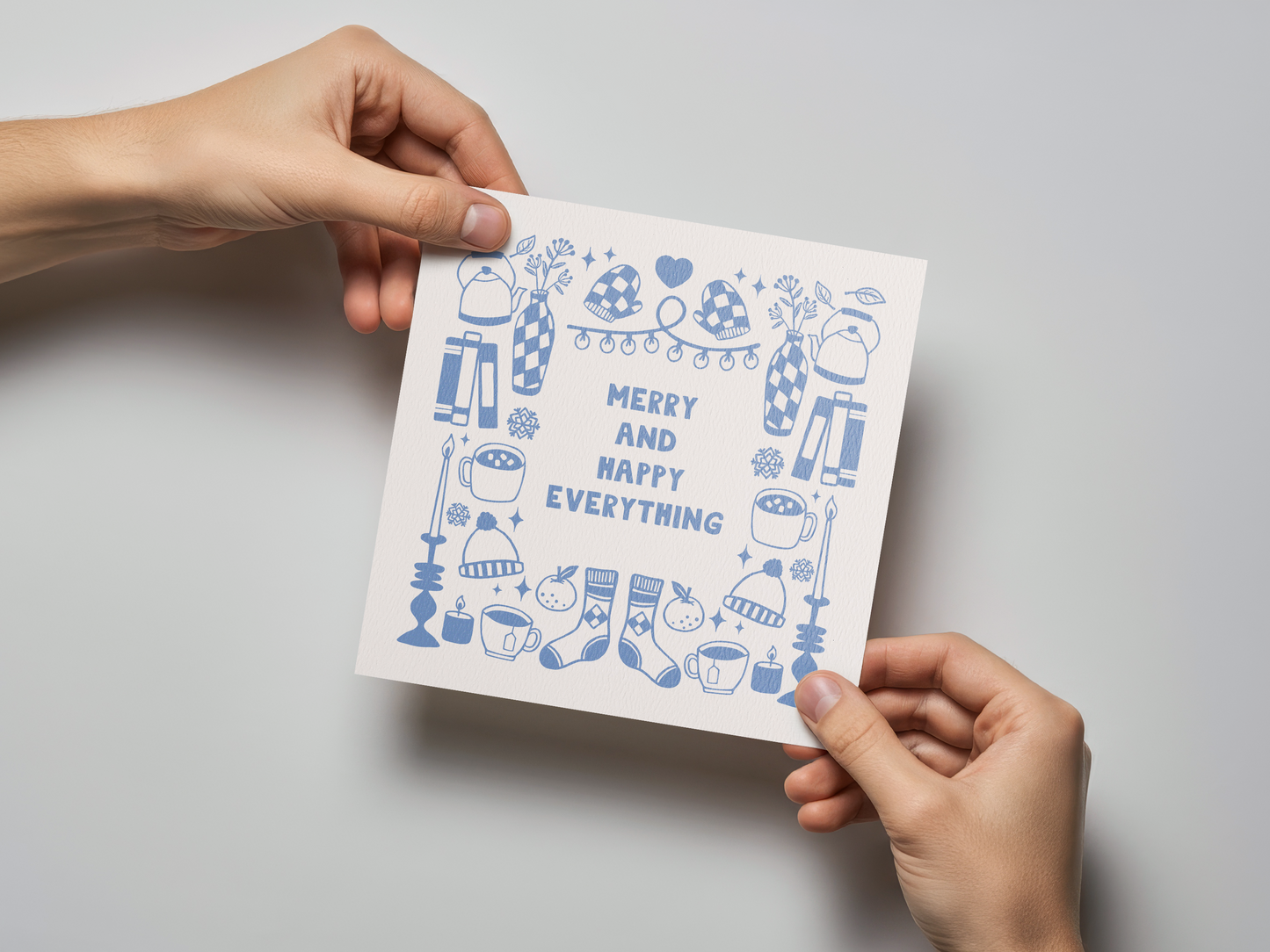 Square greeting card held by two hands featuring light blue illustrations of winter hats, mittens, candles, mugs, socks, and vases arranged around the phrase "Merry and Happy Everything" on a white background.