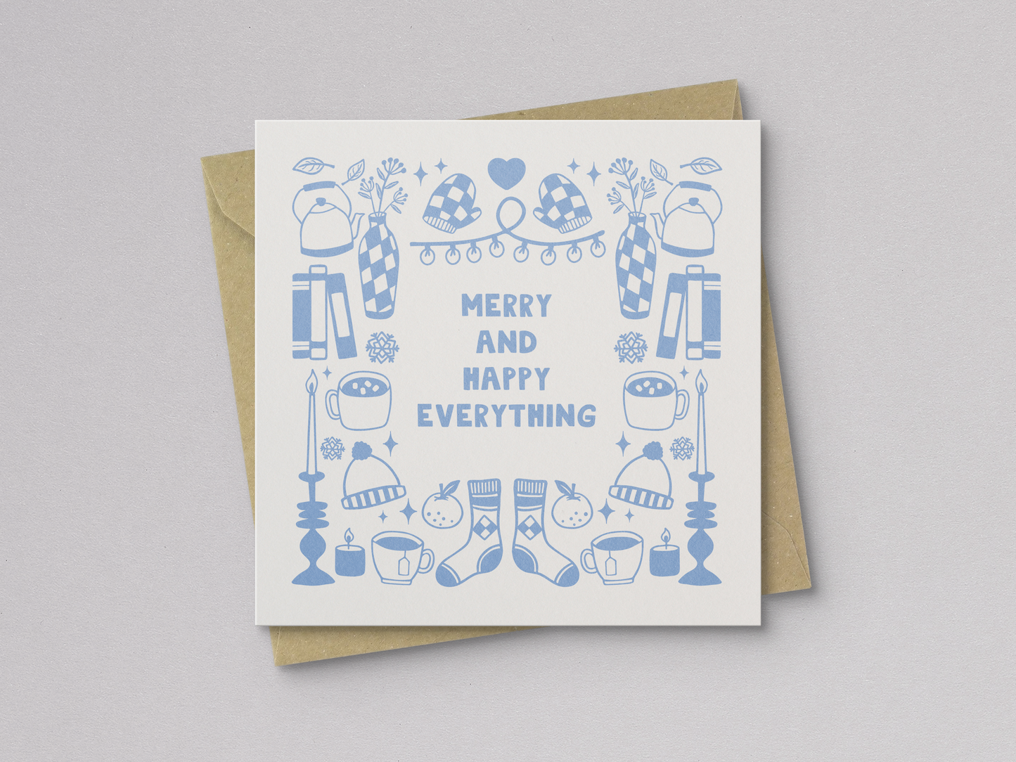 Square greeting card featuring light blue illustrations of winter-themed items including mittens, hats, socks, candles, mugs, vases, and books arranged around the text "Merry and Happy Everything," paired with a kraft paper envelope.