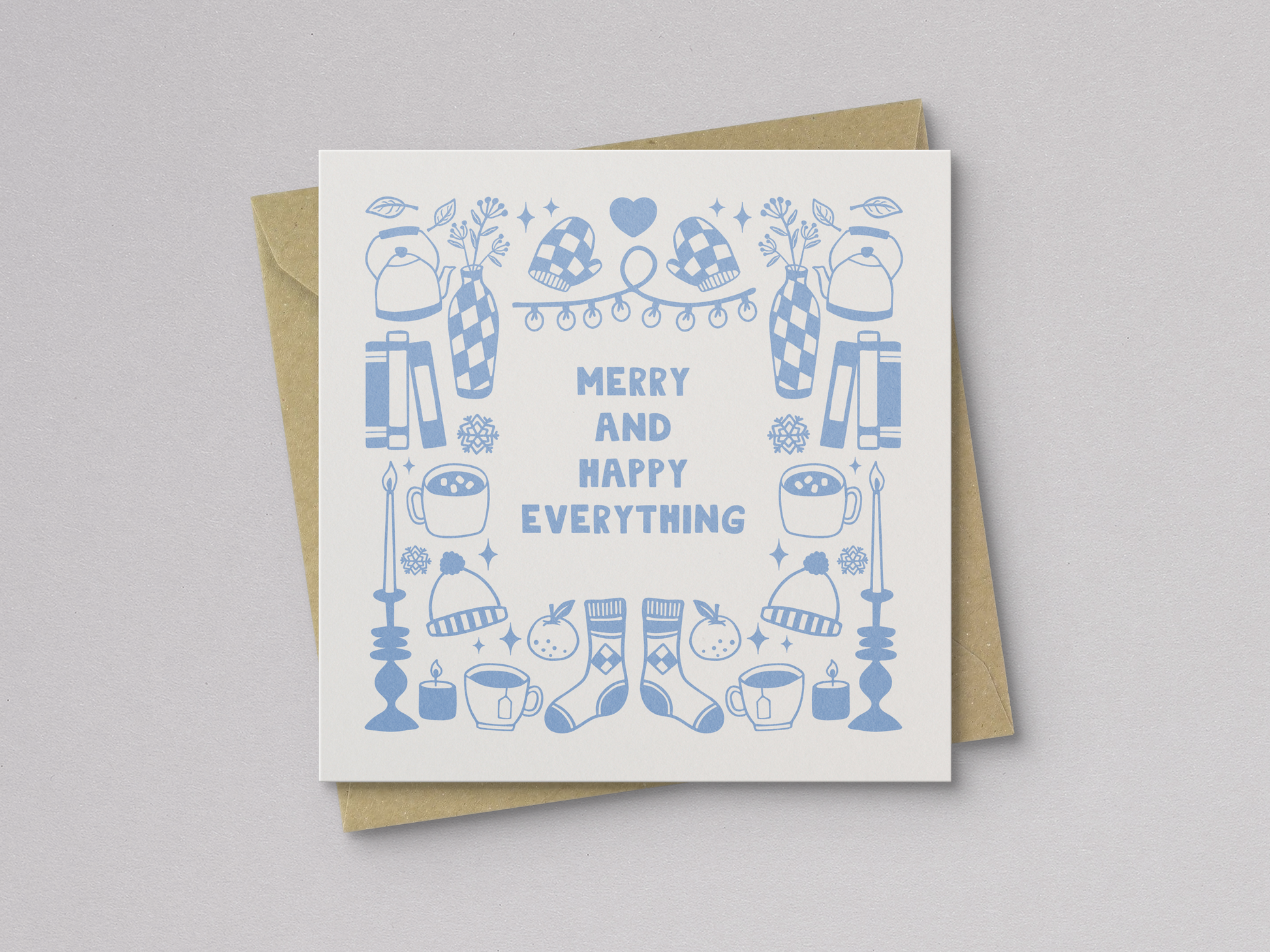 Square greeting card featuring light blue illustrations of winter-themed items including mittens, hats, socks, candles, mugs, vases, and books arranged around the text "Merry and Happy Everything," paired with a kraft paper envelope.