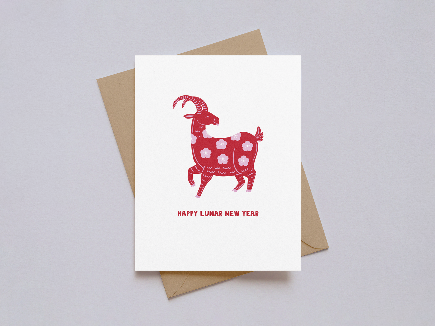 White greeting card with a red goat illustration decorated with floral patterns and the words "Happy Lunar New Year" in red, accompanied by a kraft paper envelope on a light background.