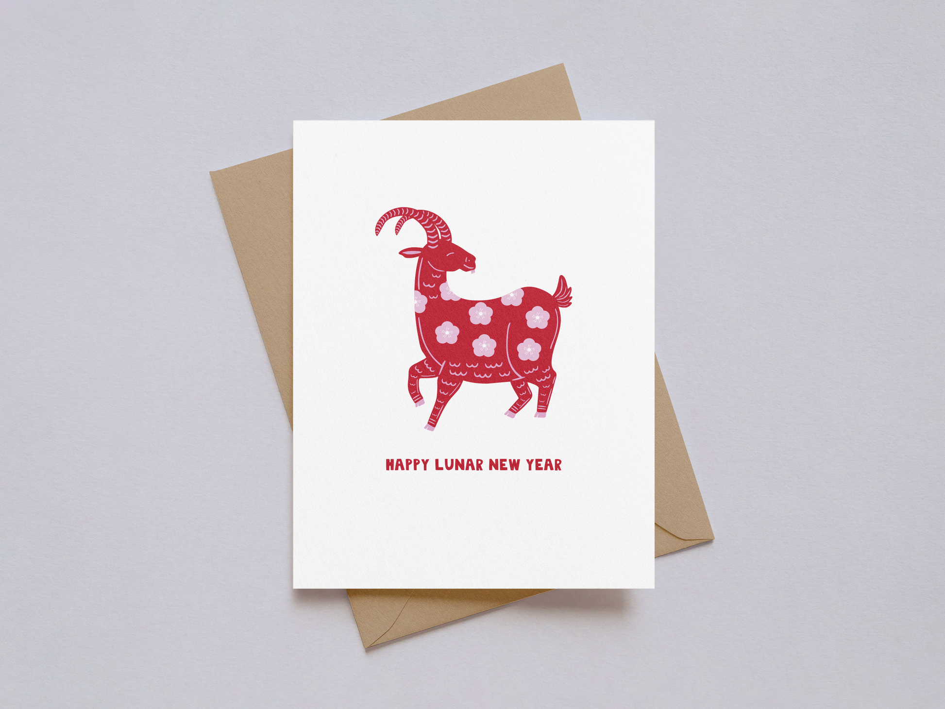 White greeting card with a red goat illustration decorated with floral patterns and the words "Happy Lunar New Year" in red, accompanied by a kraft paper envelope on a light background.
