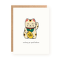 White greeting card featuring a cute hand-drawn lucky cat wearing a green bib and holding a gold coin with Chinese characters, accompanied by the message "wishing you good fortune" and a brown kraft envelope behind.