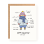 Holiday greeting card featuring a whimsical illustration of a person bundled up in multiple winter layers, labeled with various cozy clothing items like a beanie, scarf, mittens, sweaters, and boots, with the message "Happy Holidays! Stay Cozy" on a white background and accompanied by a kraft paper envelope.