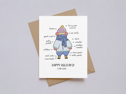 Holiday card showing a hand-drawn illustration of a person bundled in multiple layers of winter clothing including a beanie, scarf, mittens, sweaters, and boots, with labels pointing to each item and the message "Happy Holidays! Stay Cozy" on a white background, accompanied by a brown kraft paper envelope.