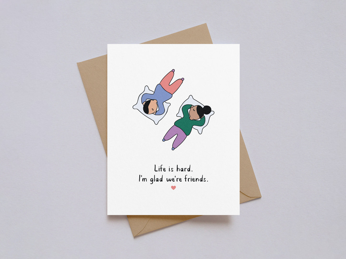 Illustrated friendship card showing two people lying on pillows with text "Life is hard. I'm glad we're friends.