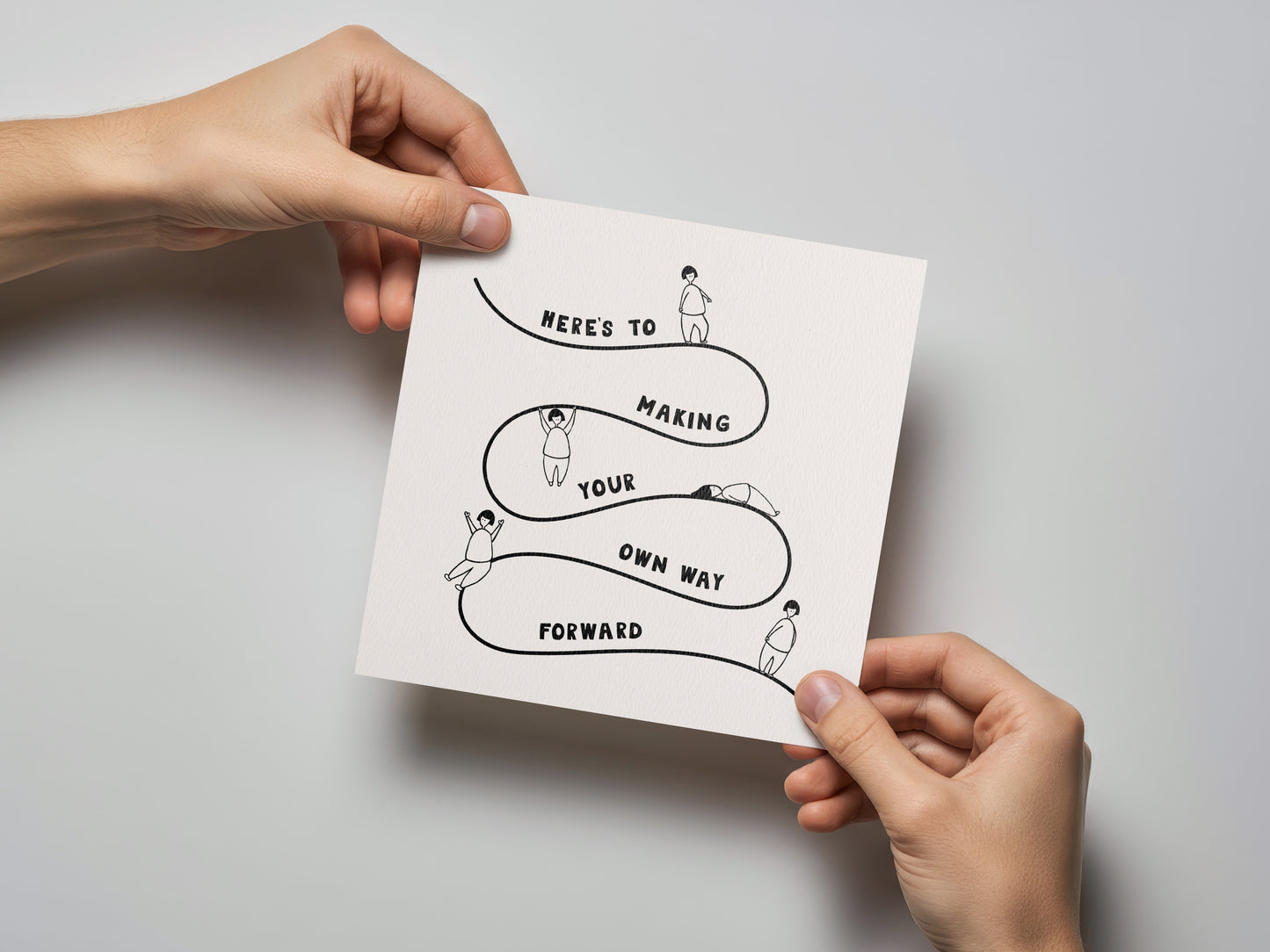 Hands holding a square encouragement card with simple black line drawings and the text "Here's to making your own way forward