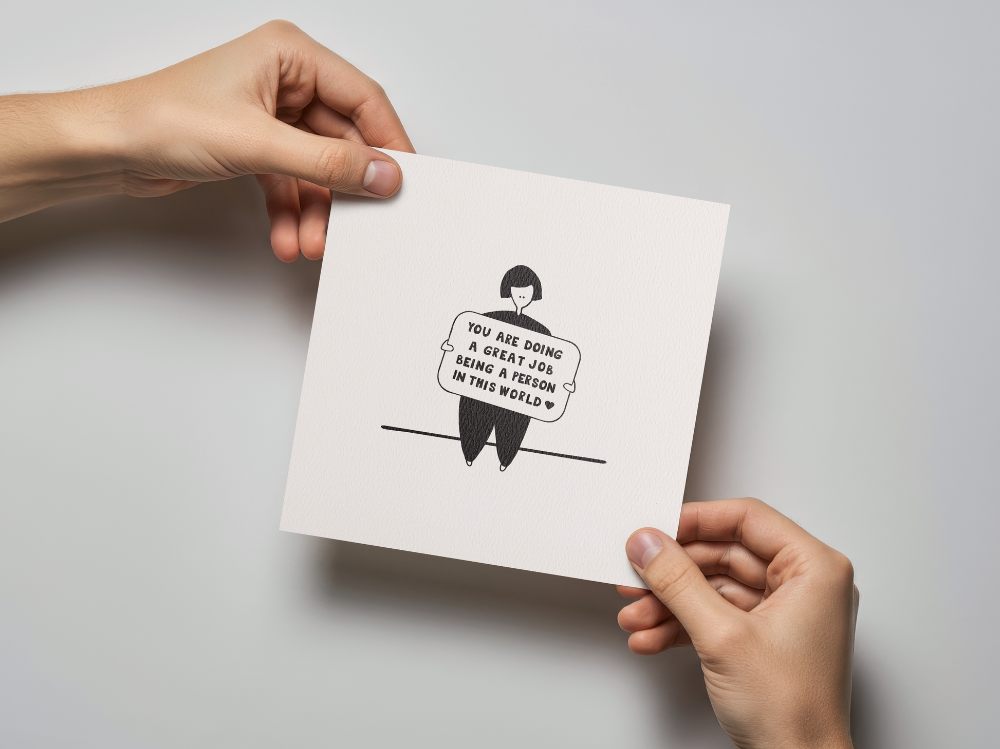 Square white greeting card held by two hands, featuring a simple black line drawing of a person sitting on a line and holding a sign with the handwritten message "You are doing a great job being a person in this world" on a textured background.