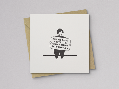 Square white greeting card featuring a simple black line drawing of a person sitting and holding a sign with the handwritten message "You are doing a great job being a person in this world," paired with a brown kraft envelope.