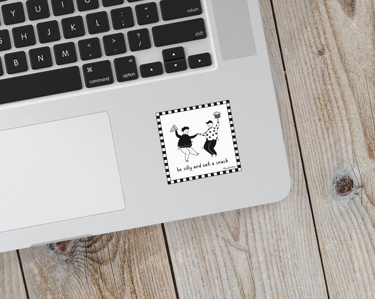 Small square sticker on a laptop showing a black and white illustration of two people dancing, one holding a slice of pizza and the other a cupcake, with the handwritten phrase "be silly and eat a snack" inside a checkered border.