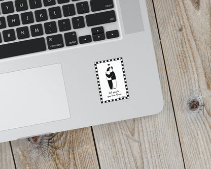 Small rectangular black and white sticker on a laptop showing an illustration of two people hugging inside a checkered border with handwritten text below saying "tell people you love them