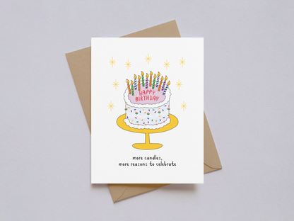 Illustrated birthday card showing a white frosted cake with colorful sprinkles and numerous lit candles on a yellow cake stand, surrounded by golden star-like sparkles and the phrase "more candles, more reasons to celebrate.