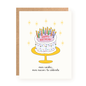 Illustrated birthday card showing a two-layer white frosted cake with colorful sprinkles and lit candles on a yellow cake stand, surrounded by golden starbursts and text reading "more candles, more reasons to celebrate.
